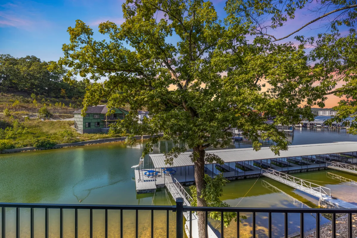 A tranquil lakeside scene featuring a large tree in the foreground, calm water, and boat docks. The view suggests accessibility to nature trails and waterfront activities, with the presence of greenery and open space that would appeal to pet owners looking for outdoor adventures.