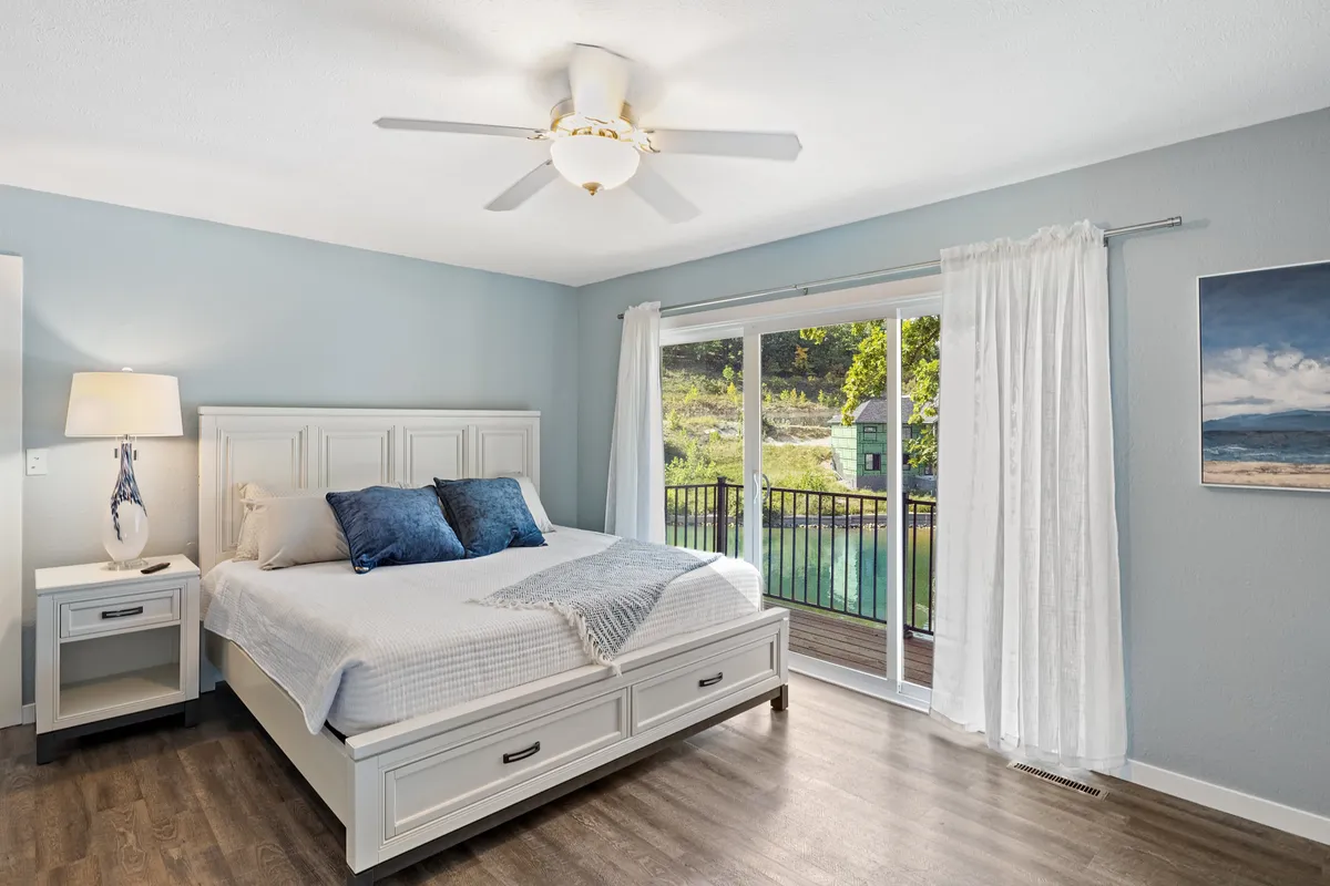 A serene bedroom featuring a bed with plush white linens and blue accent pillows, a sliding glass door that leads to a balcony, a white dresser adorned with a decorative lamp, and framed artwork on the wall. The space is bathed in natural light, enhancing the tranquil atmosphere.