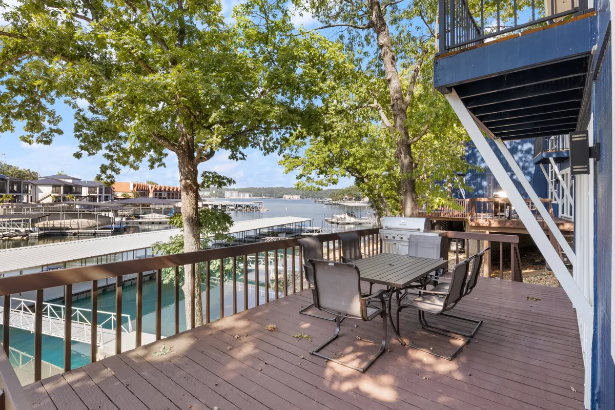 An outdoor dining table sits near the dock, which is bordered by boats and surrounded by mature trees and the sparkling lake. The setting is ideal for outdoor meals, lakeside barbecues, and watching the sunset over the water.