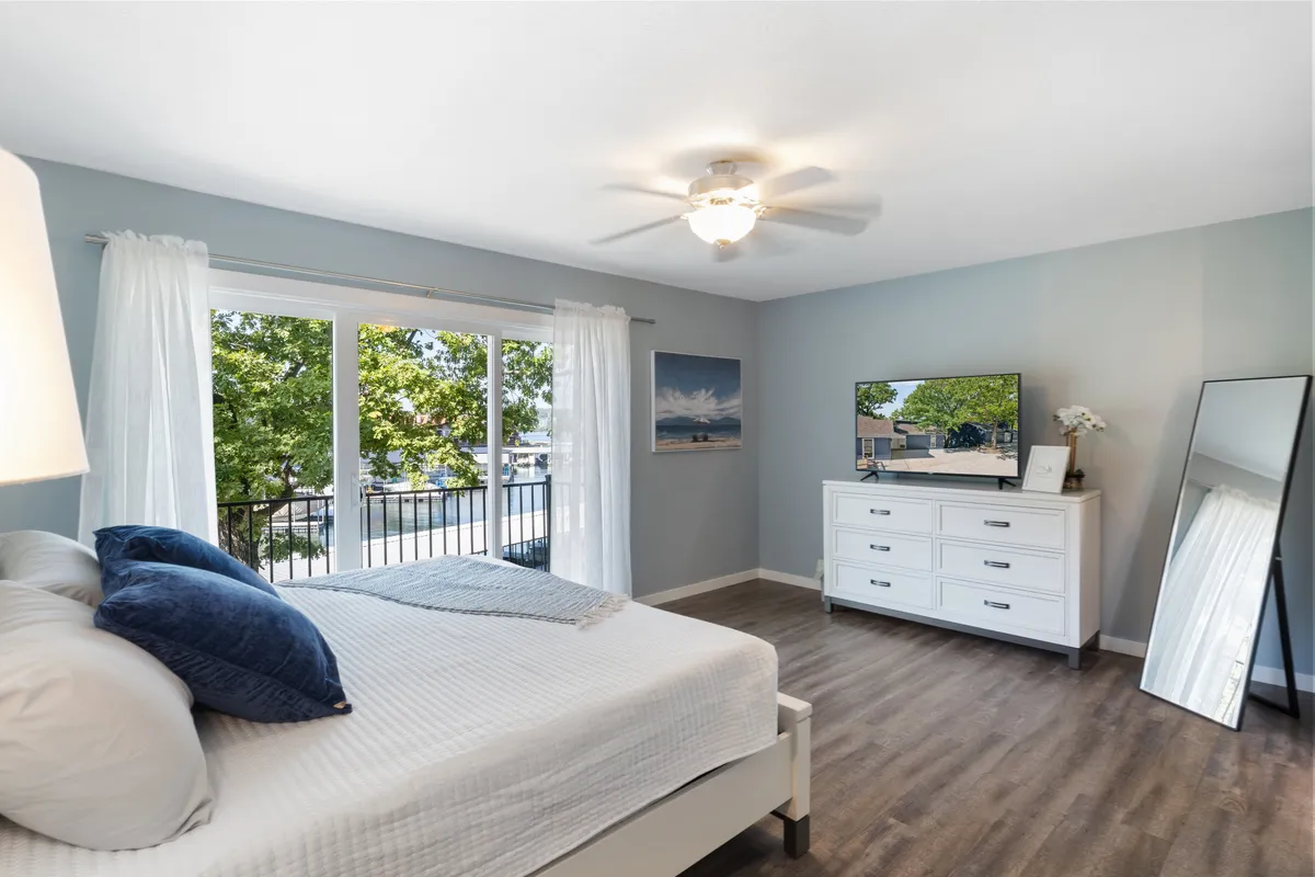 A peaceful bedroom with a neatly made bed topped by decorative pillows, large windows dressed in sheer curtains, a dresser with a mirror, and tasteful artwork on the wall. The television on the dresser and views outside add to the room's calming ambiance.