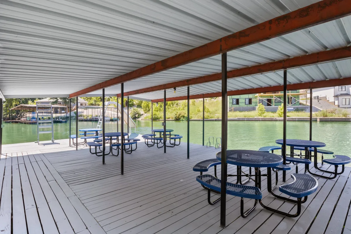 A relaxed picnic and gathering area sits at the water's edge, dotted with sturdy picnic tables and shaded by support beams, offering panoramic views of the lake. The setting feels both social and serene, suitable for both daytime and evening group activities.