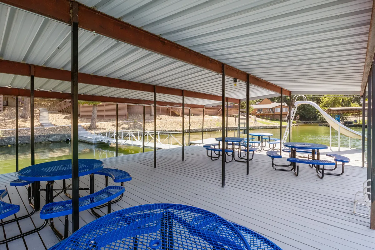 This image features blue picnic tables set near the calm lake, with a prominent water slide extending into the water and plenty of open space for families to gather. The area is surrounded by lush greenery, contributing to a fun, inviting atmosphere.