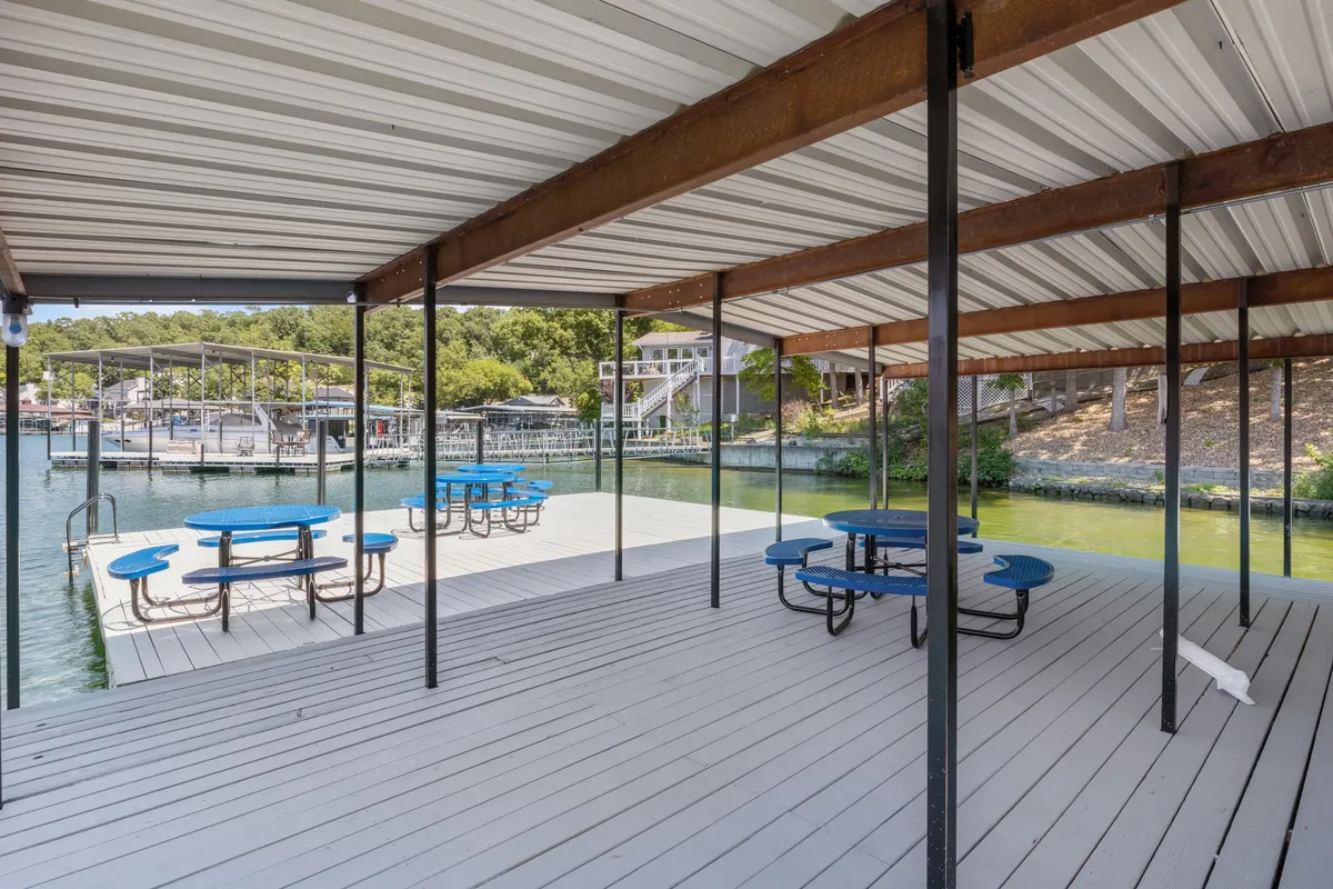A lively outdoor space with blue picnic tables, a wooden dock, surrounding greenery, and boats in the background. The atmosphere is social and casual, ideal for picnics, gatherings, and lakeside fun with pets.